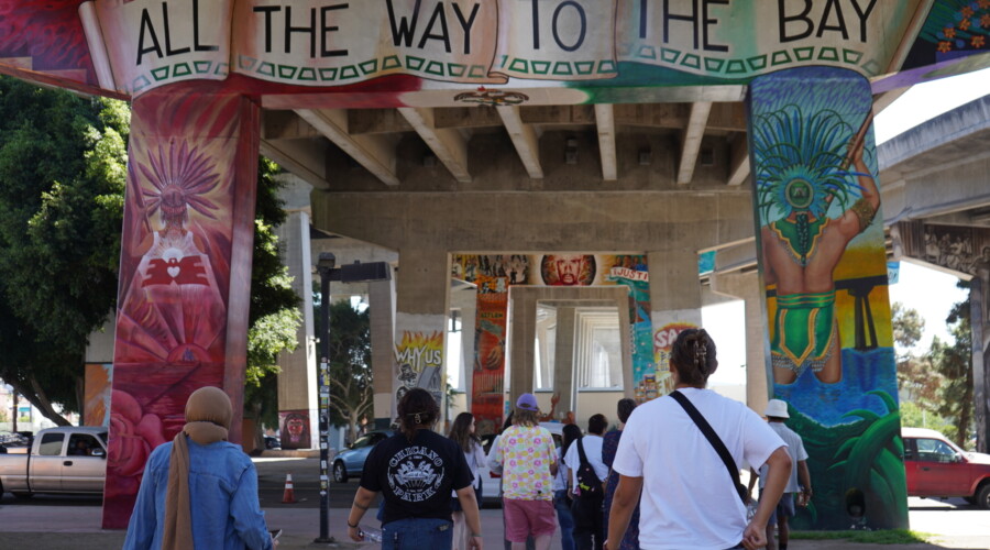A group of people in Chicano park walking under a bridge with mural art.